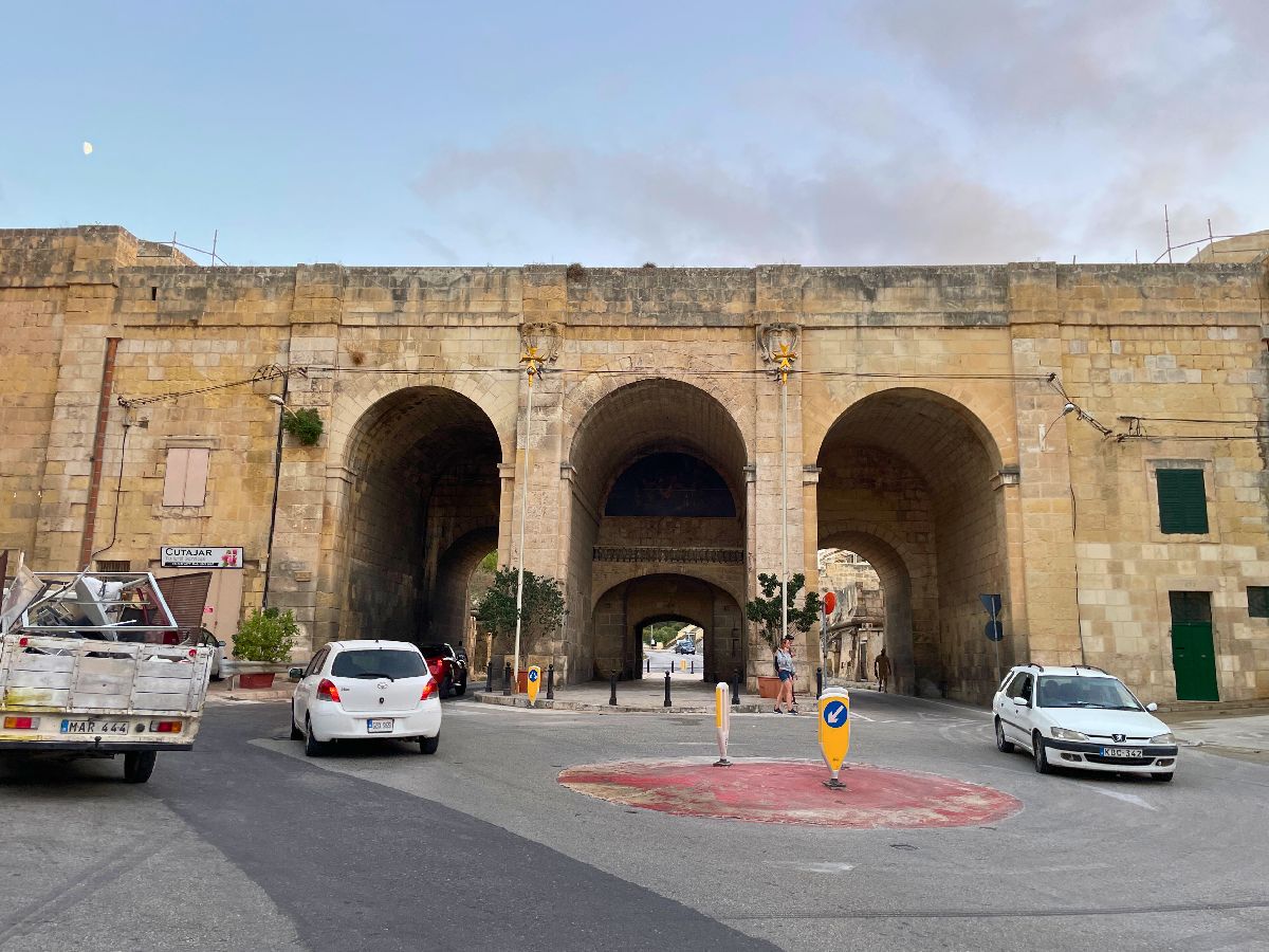 two white cars going on a small red roundabout in front of St.Helen's Gate in Cospicua